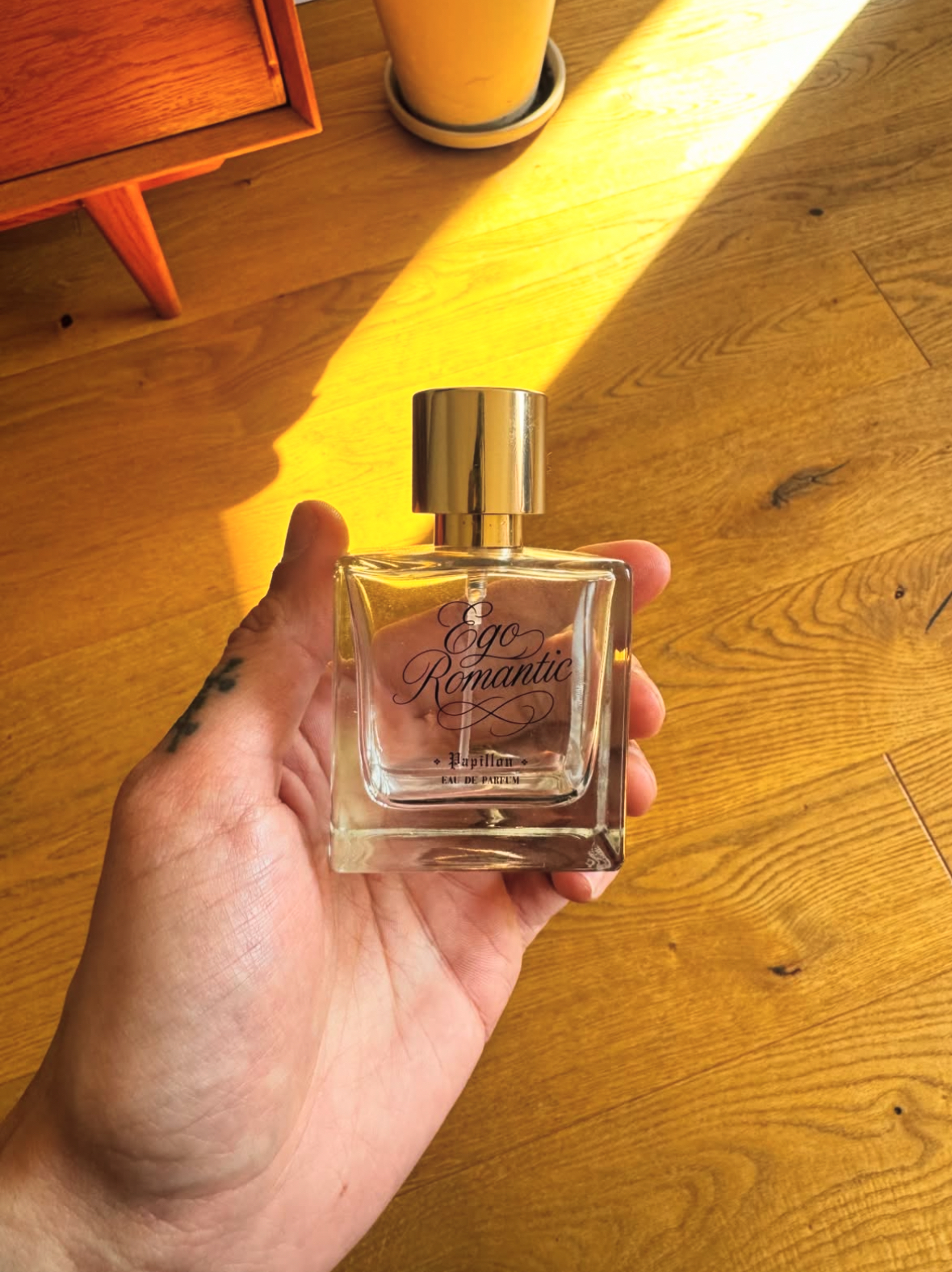 Hand holding a square Ego Romantic perfume bottle in warm sunlight on a wooden floor